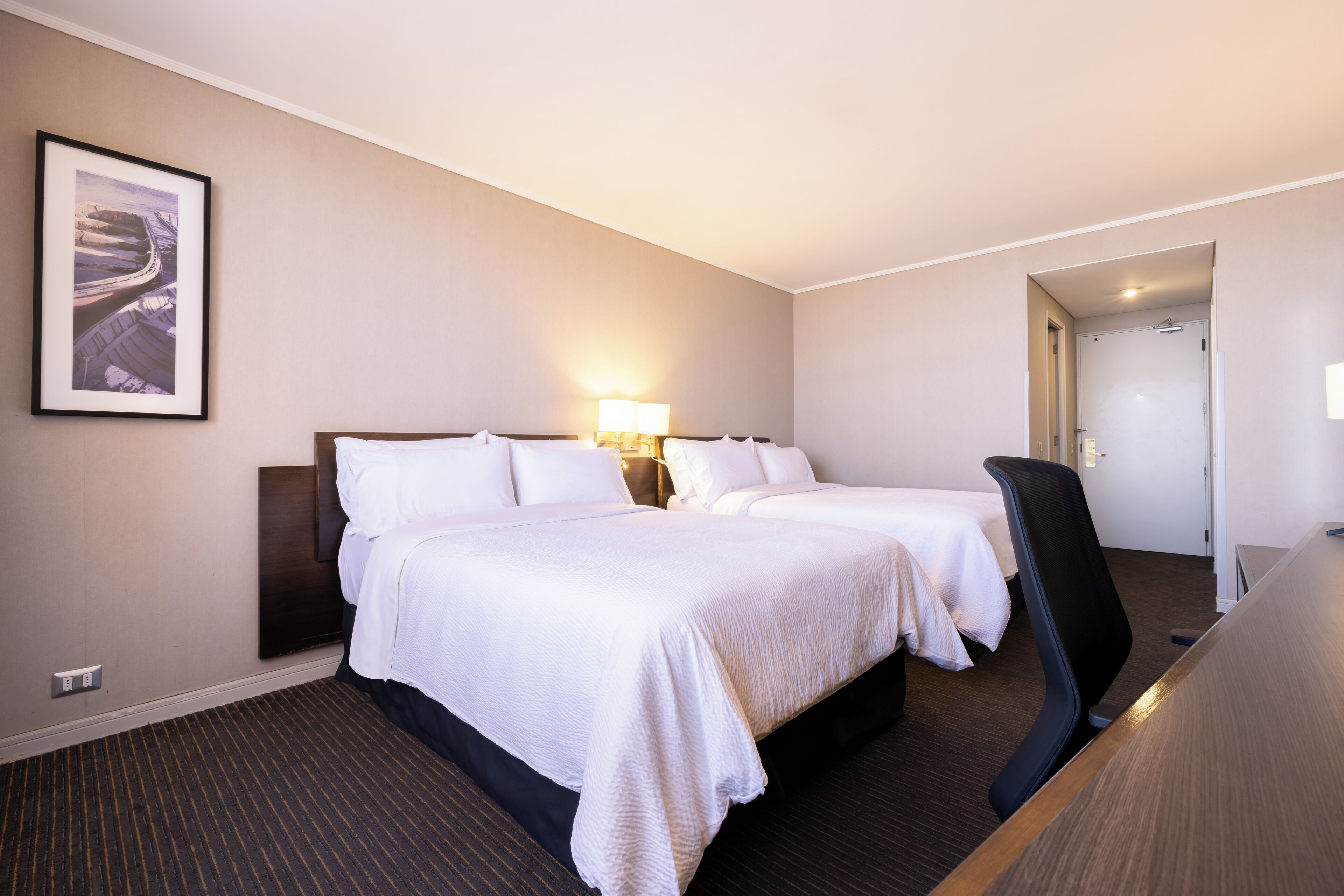 A hotel room with two beds, a desk, a chair, a lamp, and a framed picture on the wall.