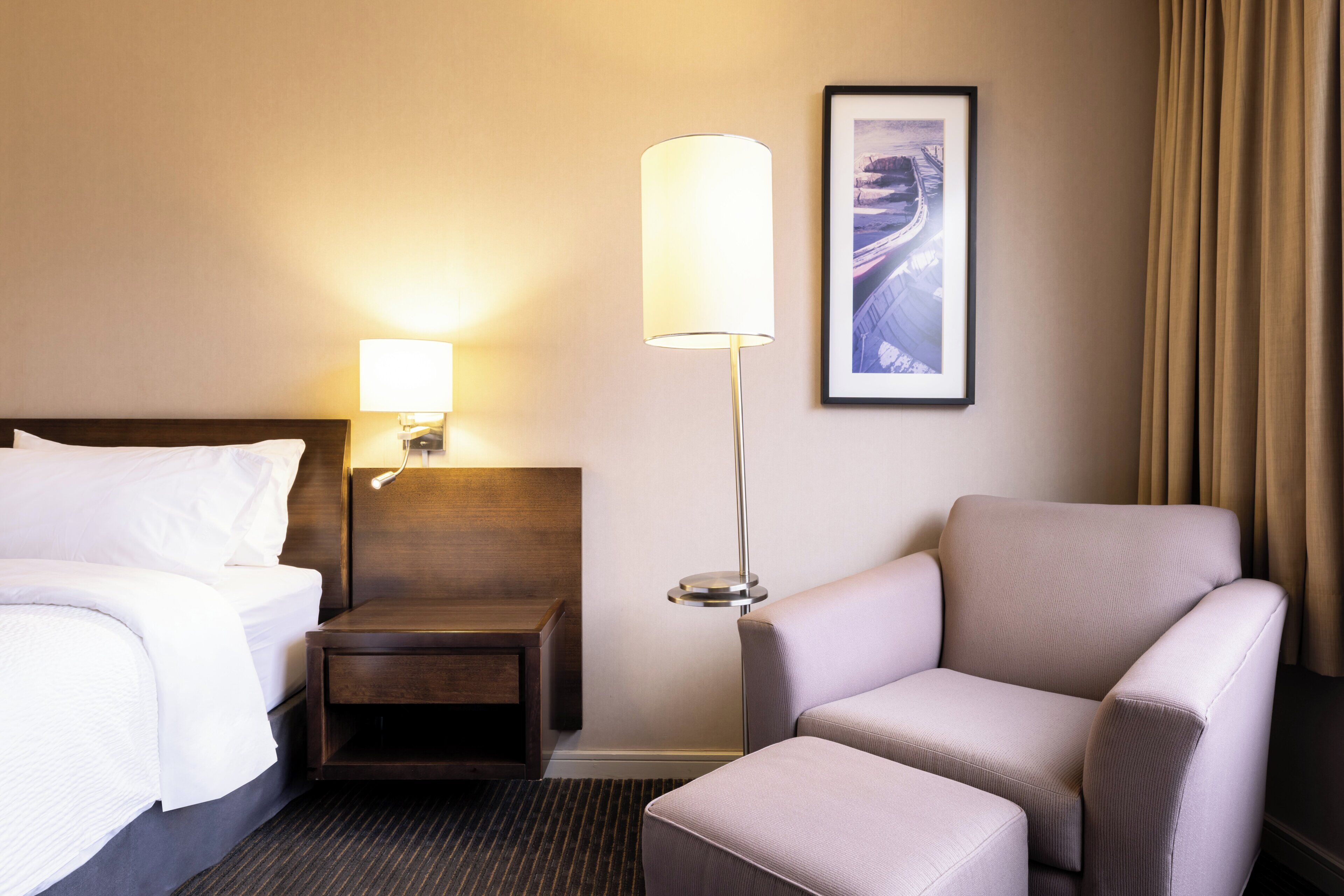 A hotel room with a bed, a nightstand, a lamp, a chair, and a framed picture on the wall.