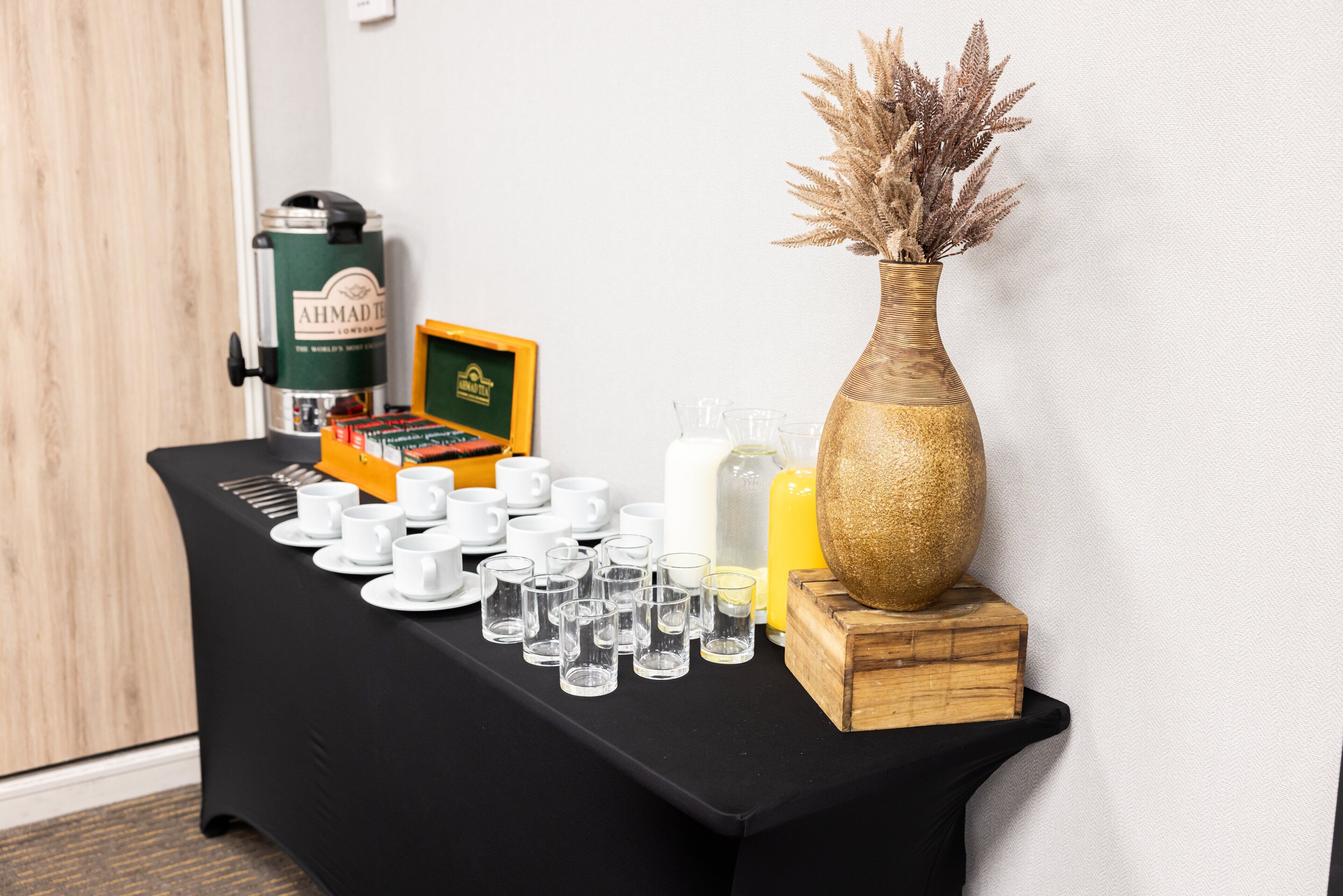 A table with a coffee maker, a box of tea, and a vase with dried plants.