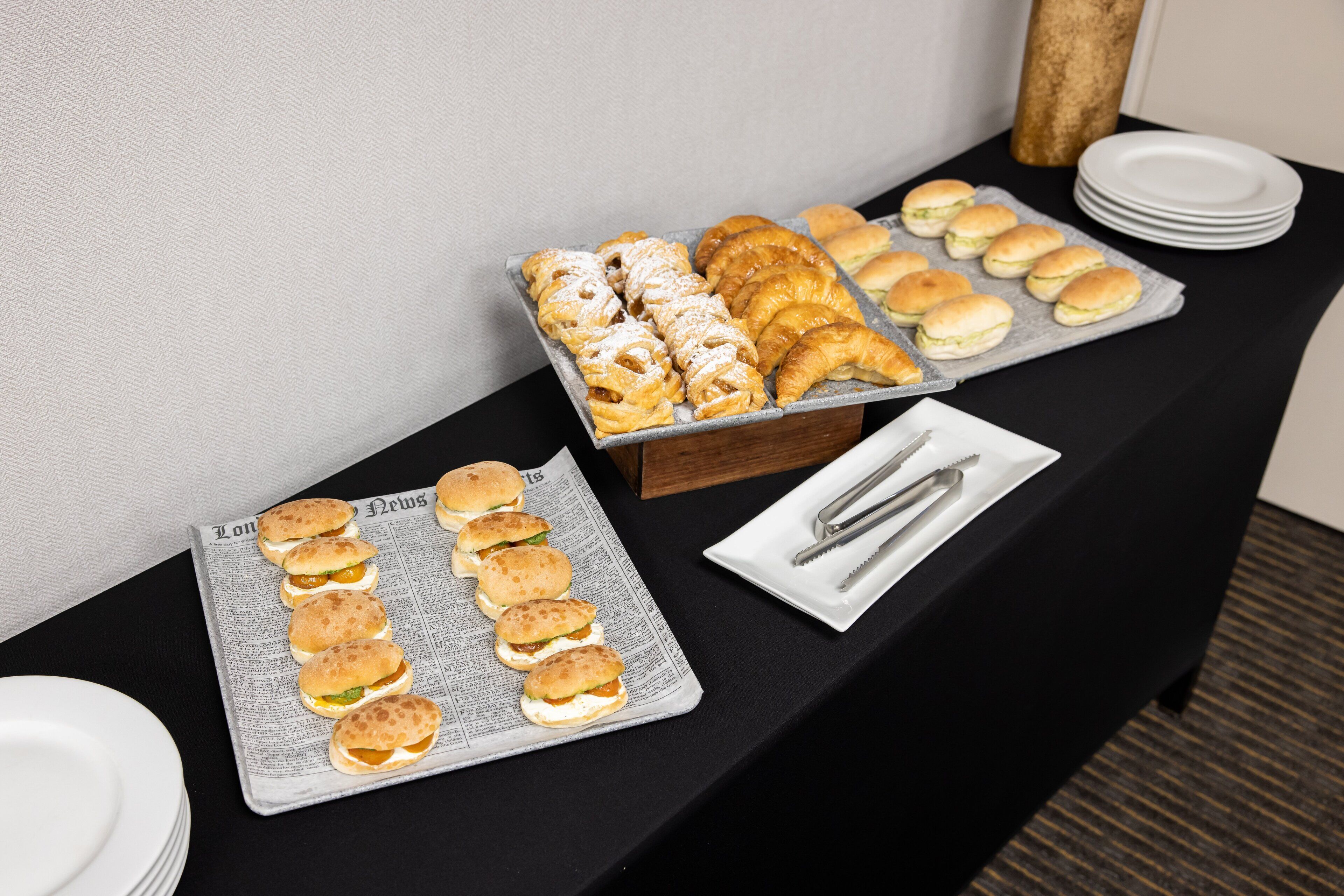 A buffet table with various pastries and sandwiches, plates, and cutlery.