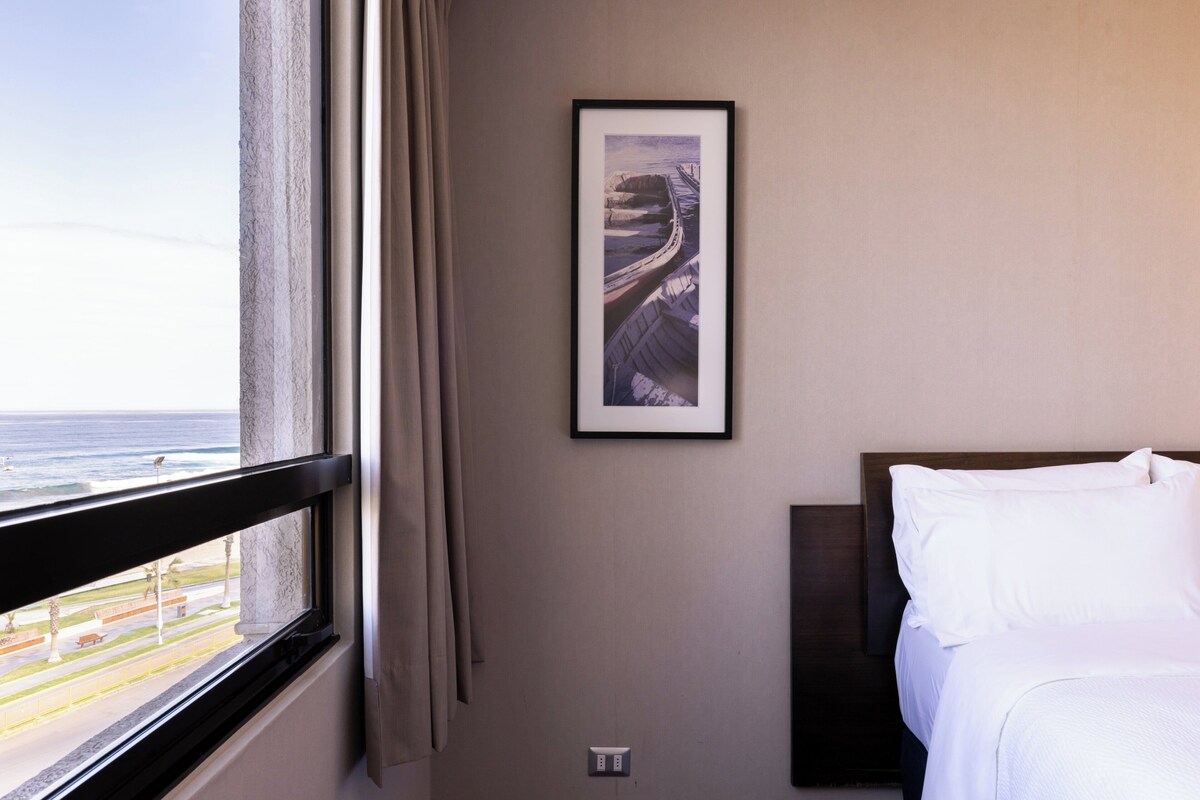 A hotel room with a bed, a framed picture on the wall, and a window with a view of the ocean.