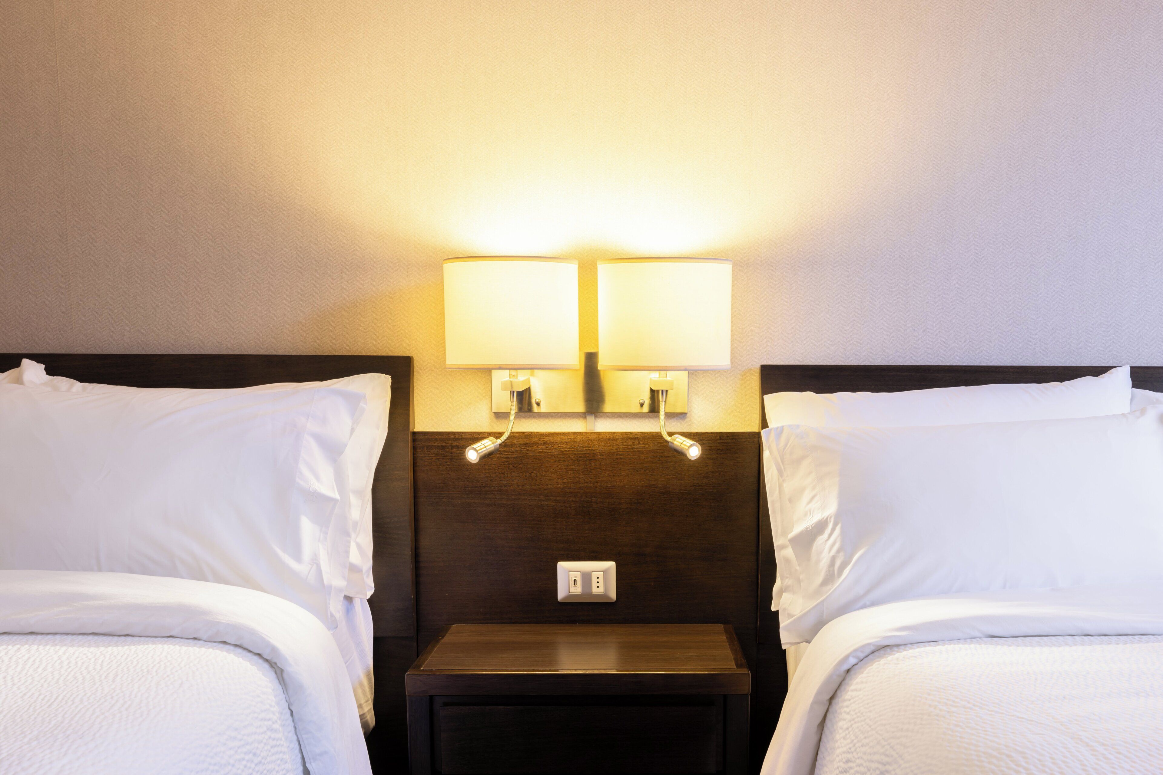 A hotel room with two beds, a nightstand, and two wall-mounted lamps.