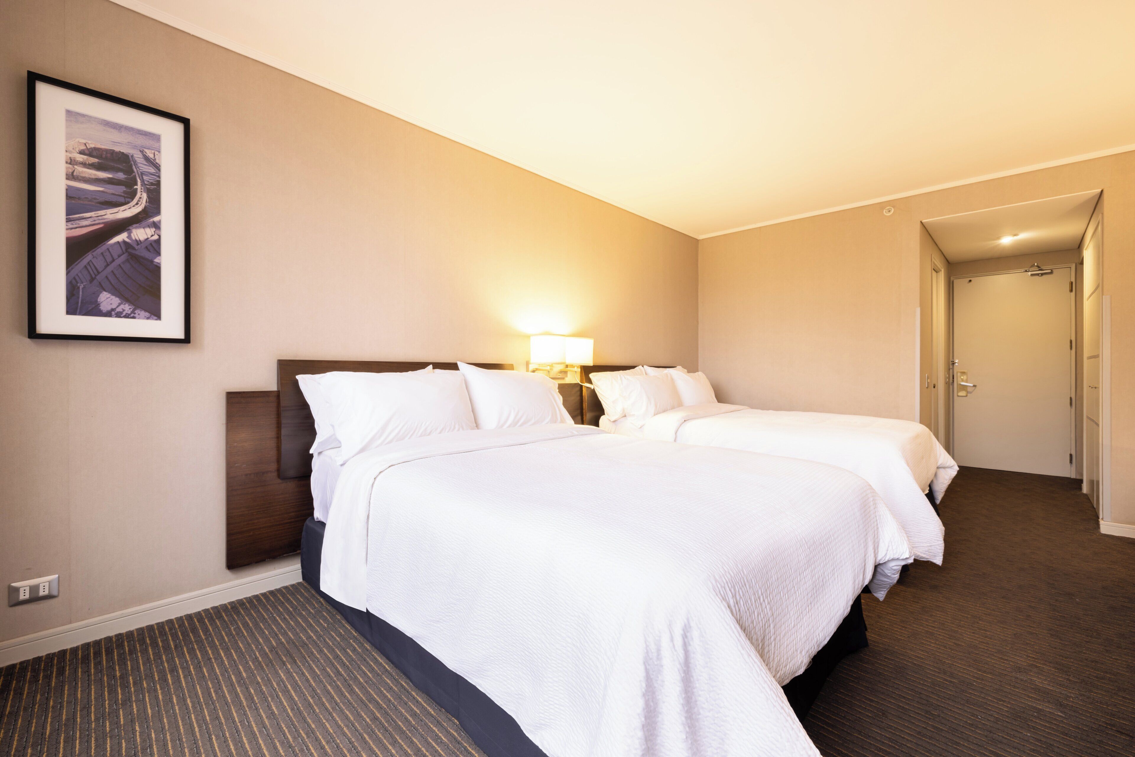 A hotel room with two beds, a nightstand, a lamp, a framed picture on the wall, and a carpeted floor.