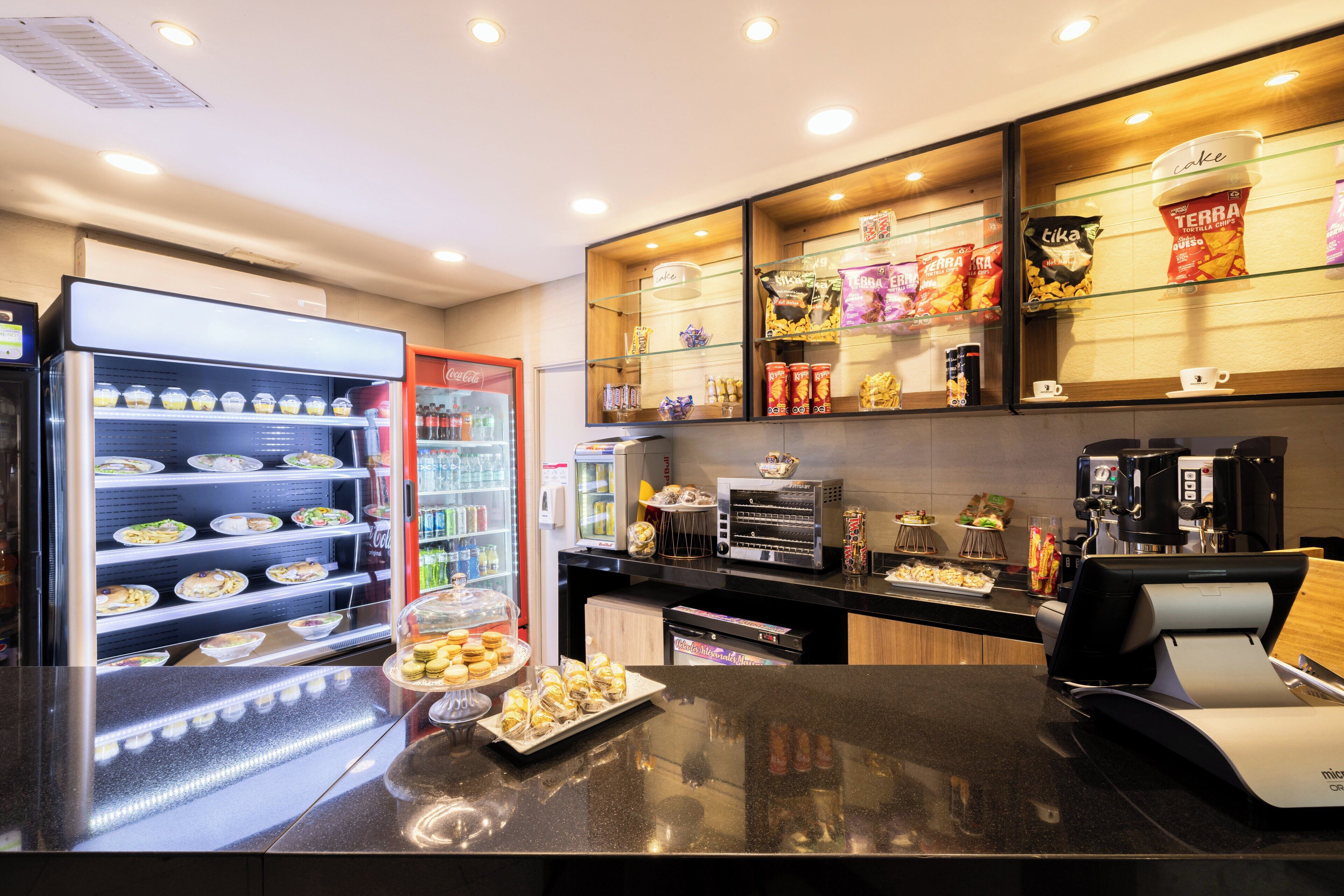 Een modern café met een bedieningsluik, een koelvitrine met etenswaren en diverse snacks en dranken op planken.
