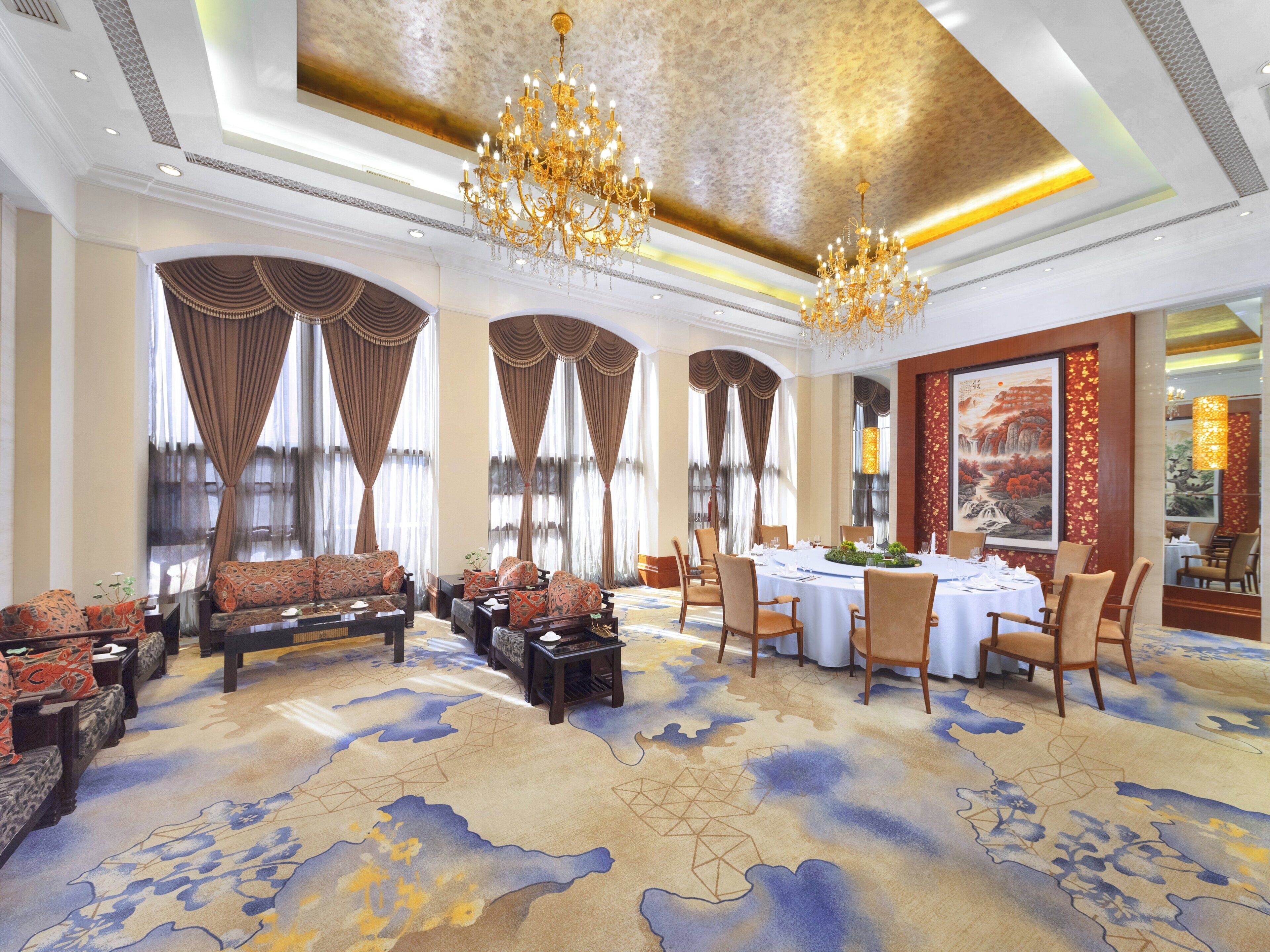 Een ruime hotel lobby met een groot eetgedeelte, elegante kroonluchters en een vloerkleed met een blauw en gouden ontwerp.