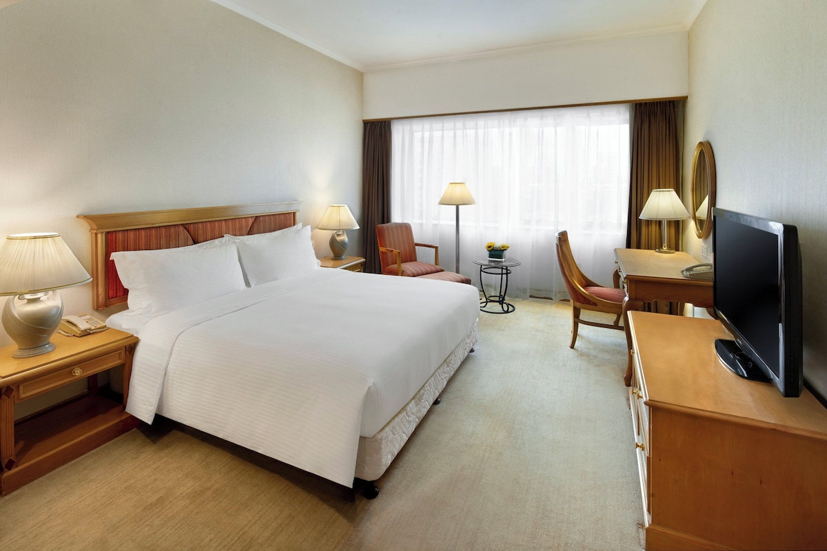 A hotel room with a large bed, a desk with a TV, a chair, two lamps, and a small table with flowers.