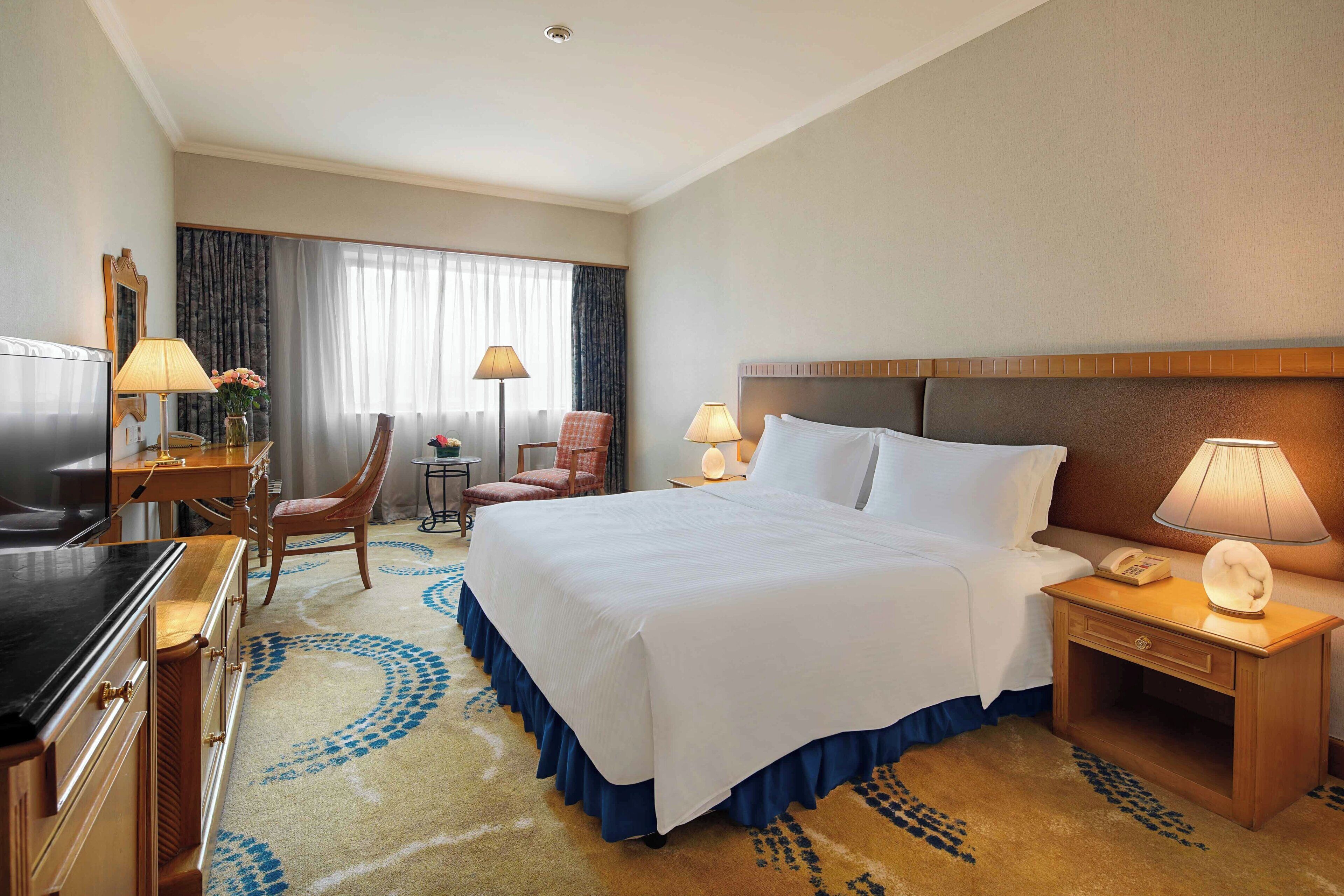 A hotel room with a bed, two bedside tables, a desk with a chair, a TV, and a large window with curtains.