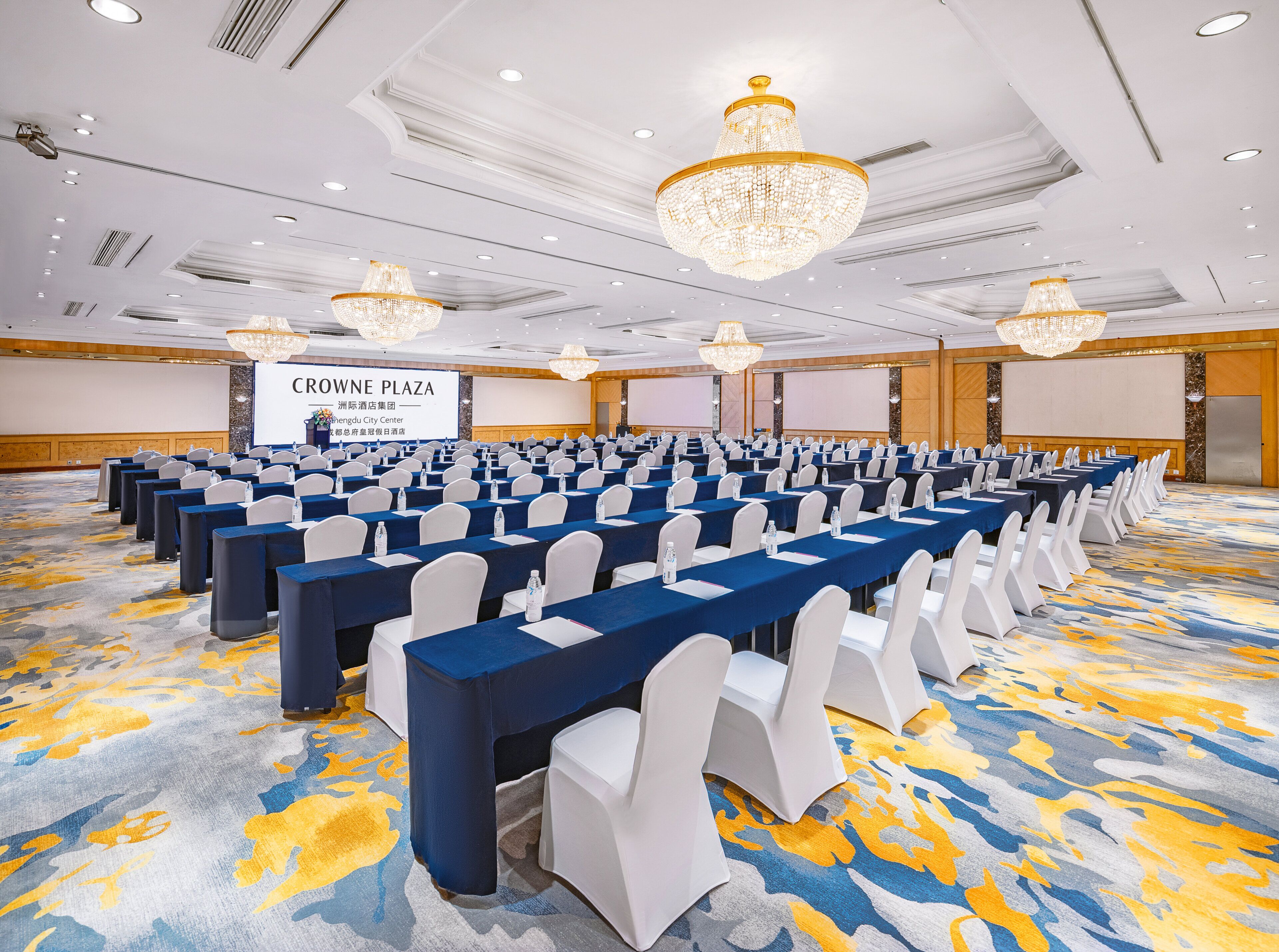 A conference room with rows of tables, white chairs, and a large screen.