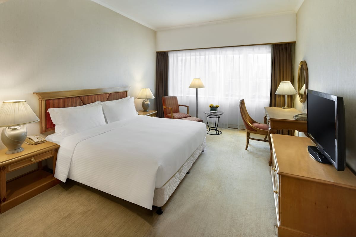 A hotel room with a large bed, a desk with a TV, a chair, two lamps, and a small table with flowers.