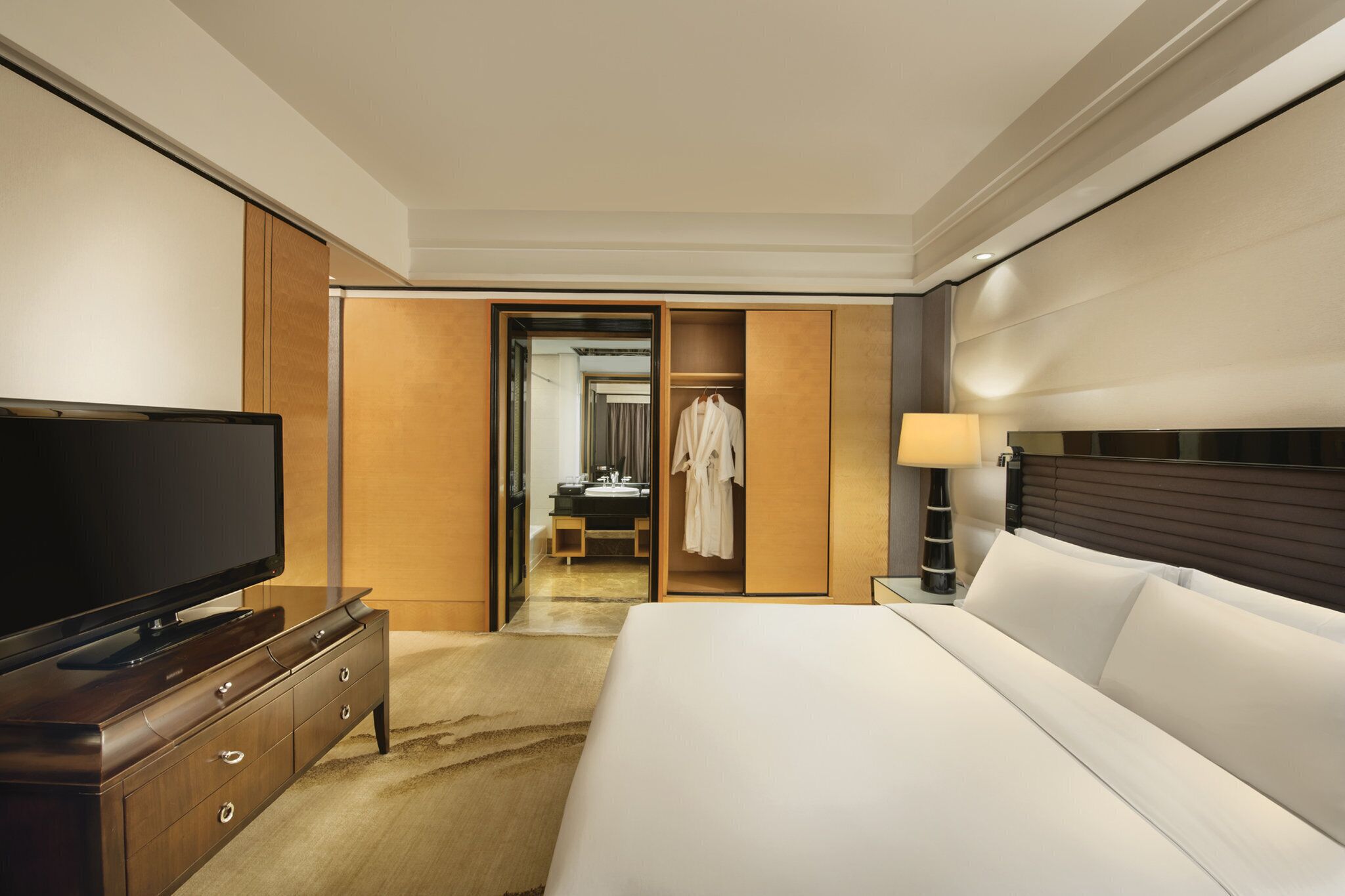 A hotel room with a large bed, a flat-screen TV on a wooden dresser, a bedside table with a lamp, and a walk-in closet with a robe hanging.