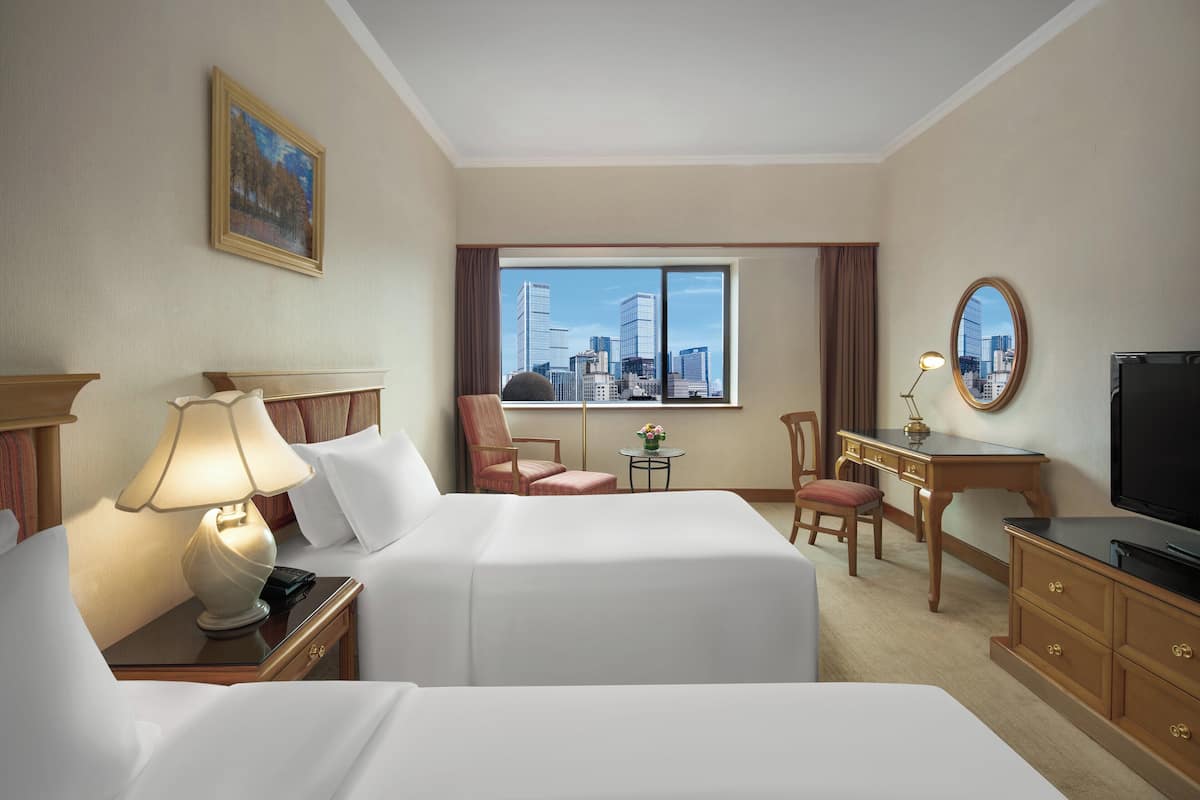 A hotel room with two beds, a desk, a chair, a TV, and a large window with a city view.
