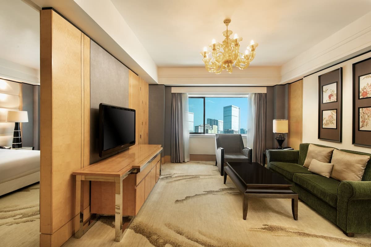 A modern hotel room with a flat-screen TV, a wooden desk, a green sofa, a chandelier, and a city view.