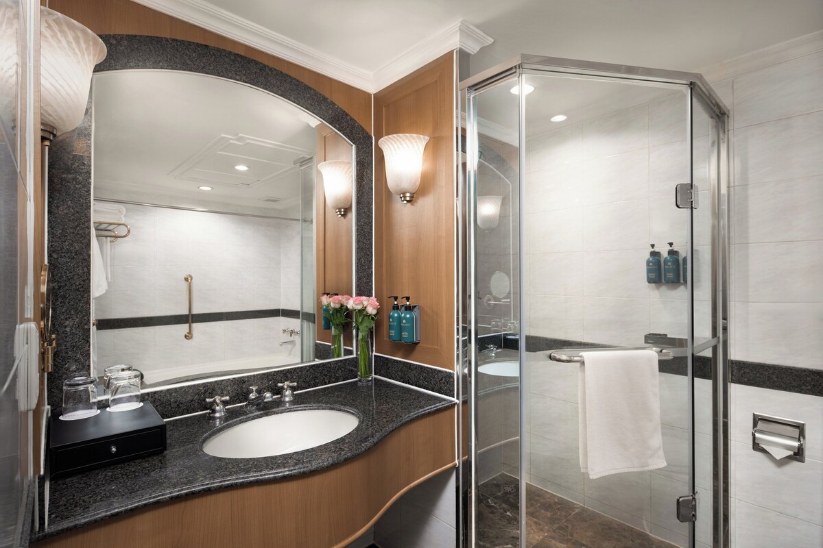 A modern bathroom with a granite countertop, a large mirror, a glass shower enclosure, and a white towel hanging on the shower door.