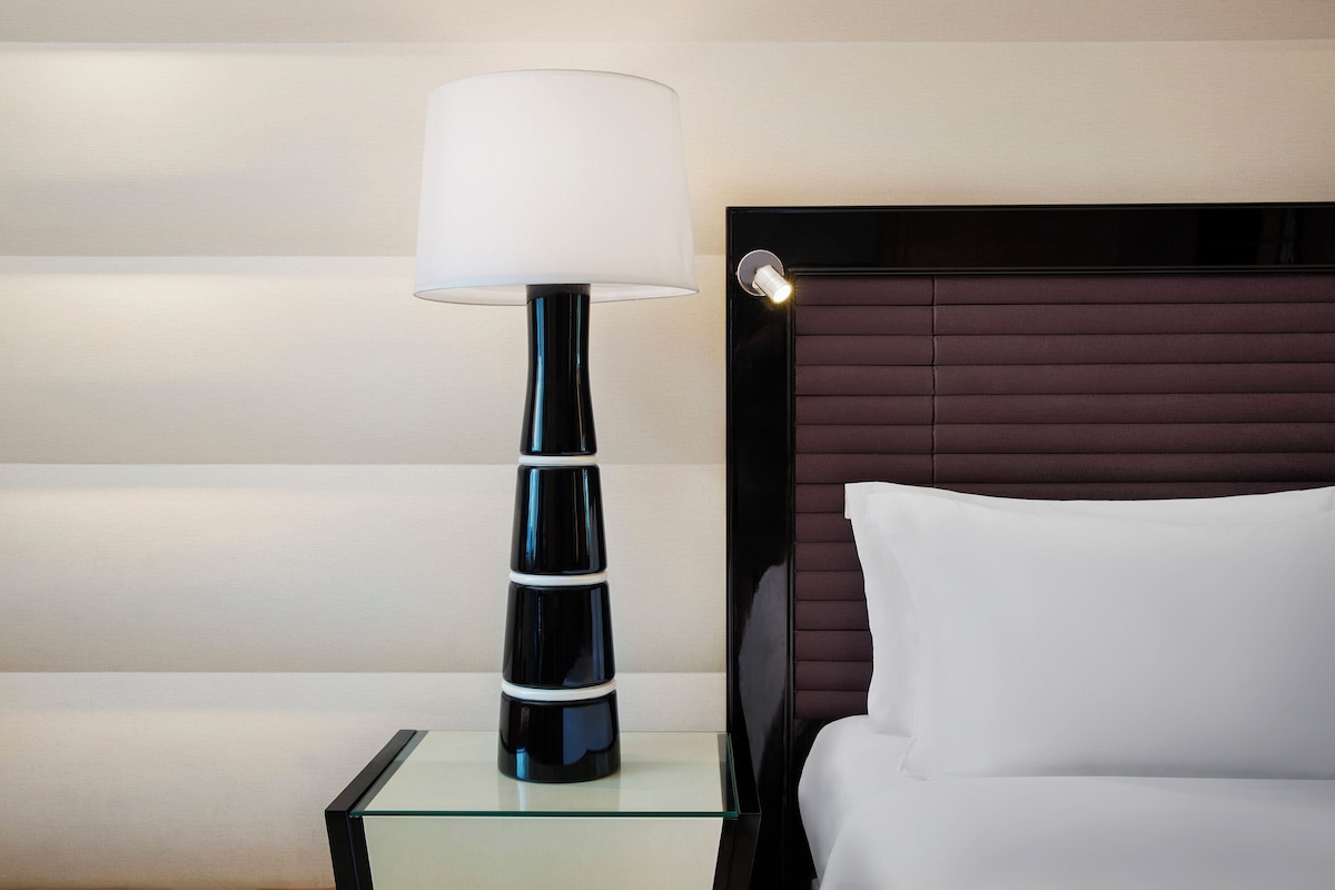 A modern hotel room with a glass side table, a black lamp with a white shade, a headboard with horizontal lines, and a white pillow.