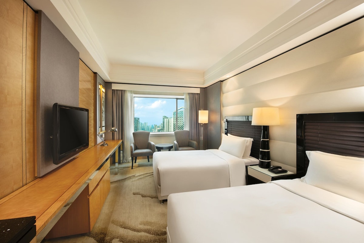 A hotel room with two beds, a TV, a desk, and a view of the city.