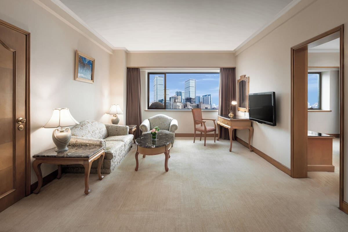 A hotel room with a city view, a sofa, a coffee table, a desk, and a television.