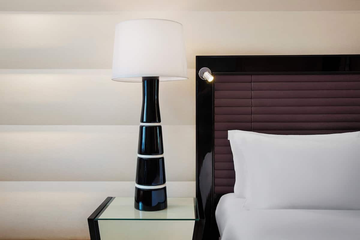 A modern hotel room with a glass side table, a black lamp with a white shade, a headboard with horizontal lines, and a white pillow.