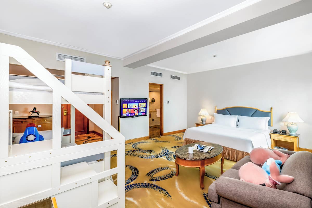 A hotel room with a bunk bed, a television, a bed with a blue headboard, a nightstand with a lamp, and a carpet with a circular pattern.