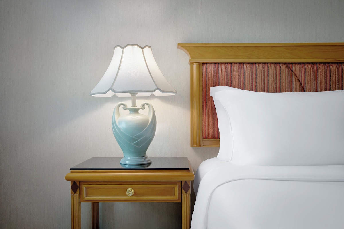 A neatly made bed with a wooden headboard and a striped curtain, a small bedside table with a lamp, and a drawer with a gold knob.