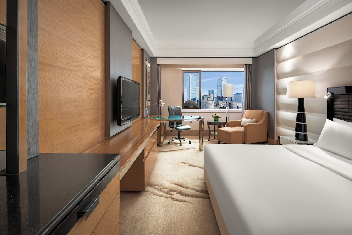 A modern hotel room with a large bed, a desk with a flat-screen TV, a comfortable armchair, a small table with a lamp, and a view of the city skyline through large windows.
