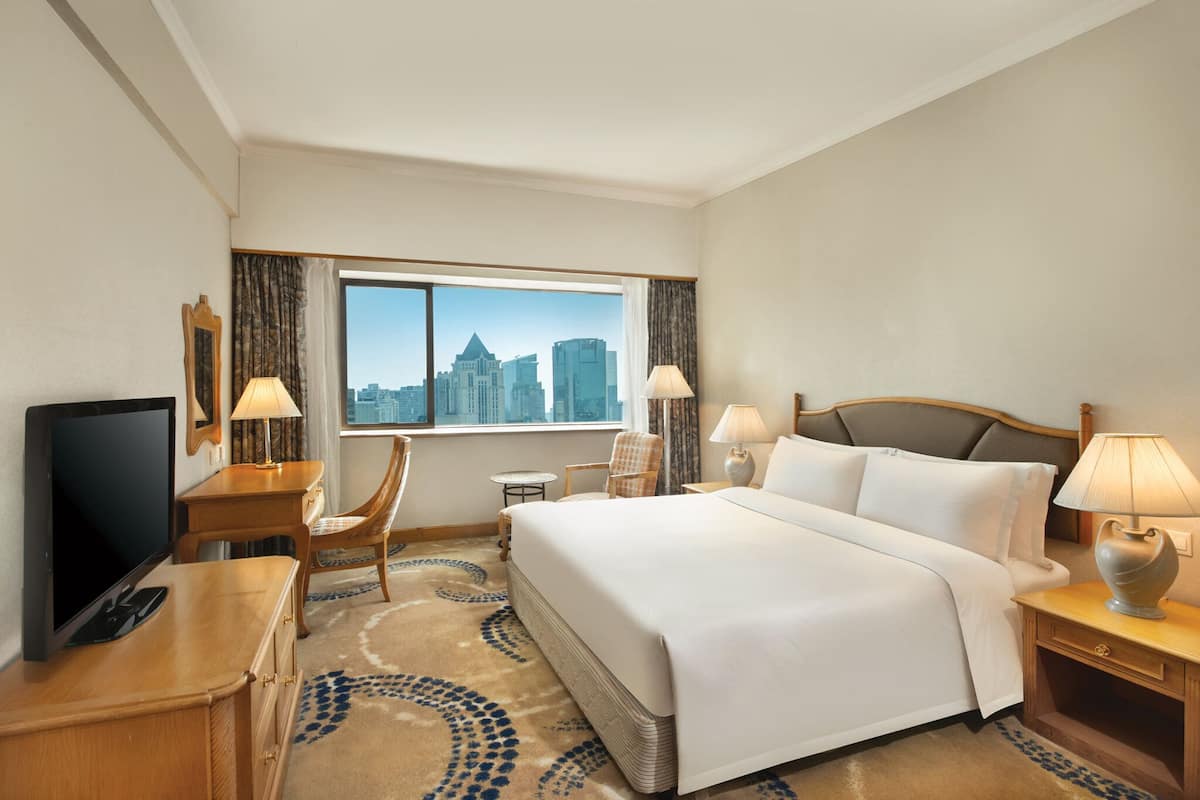 A hotel room with a large bed, a desk with a TV, two chairs, a small table, and a view of a city skyline through a large window.