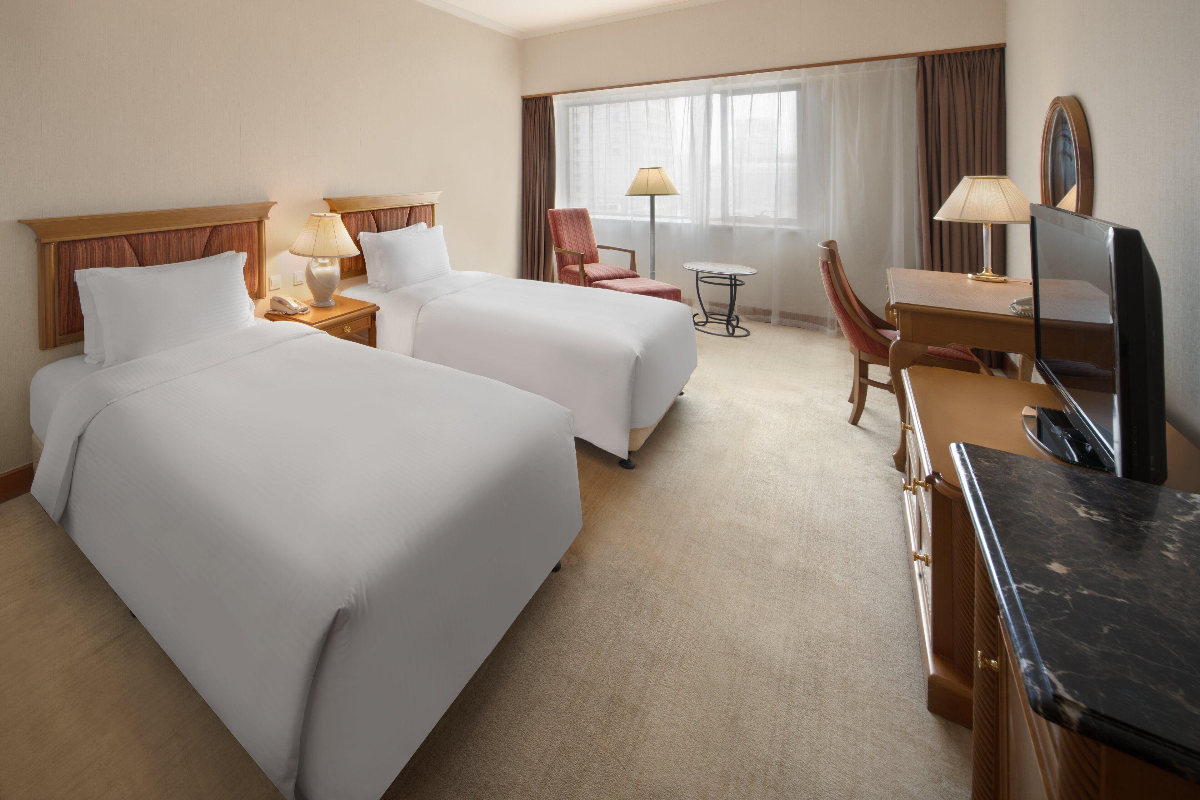 A hotel room with two beds, a desk, a chair, a TV, and a small table with lamps.