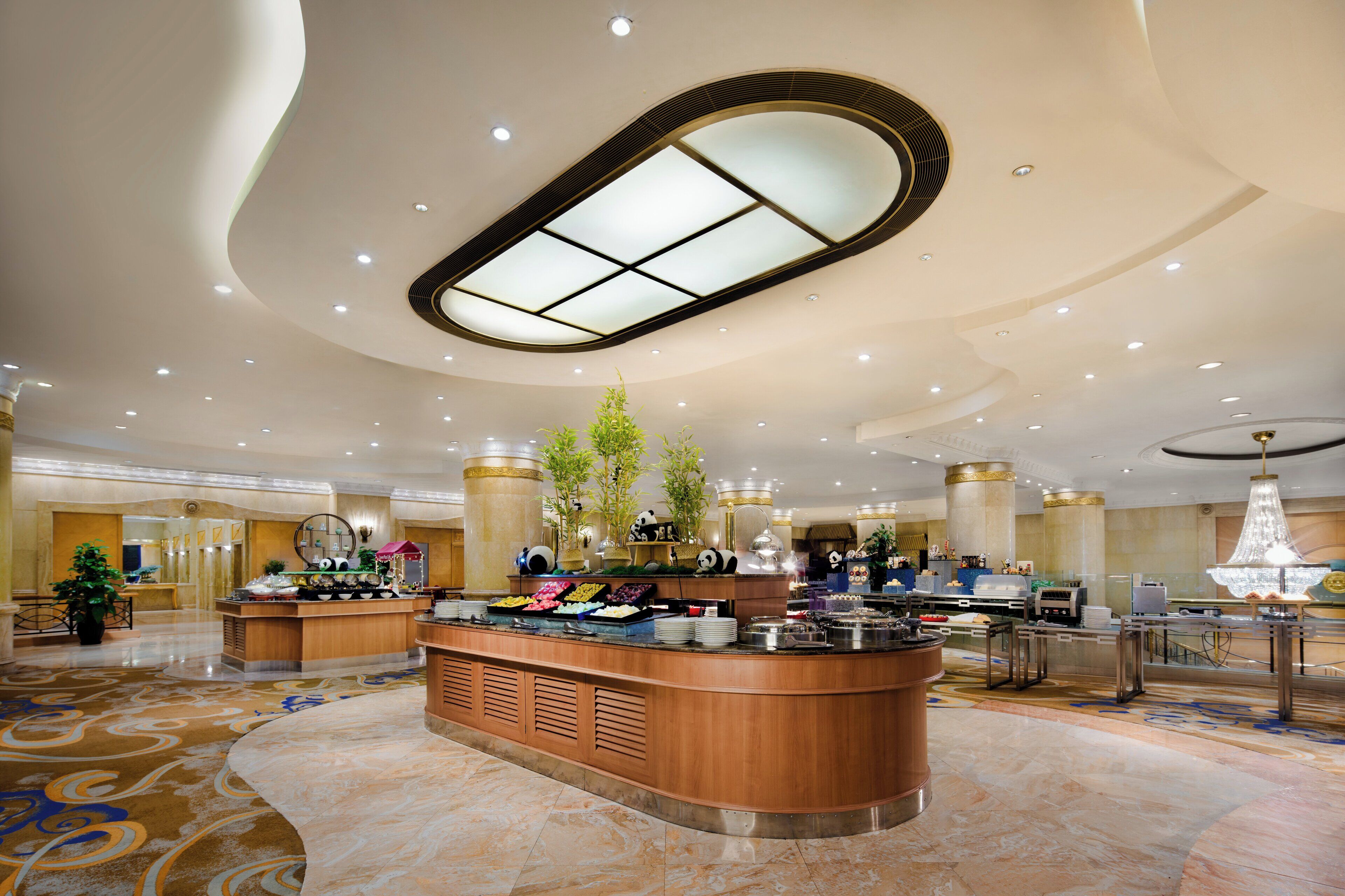 A spacious hotel lobby with a central reception desk, a large chandelier, and a decorative ceiling design.