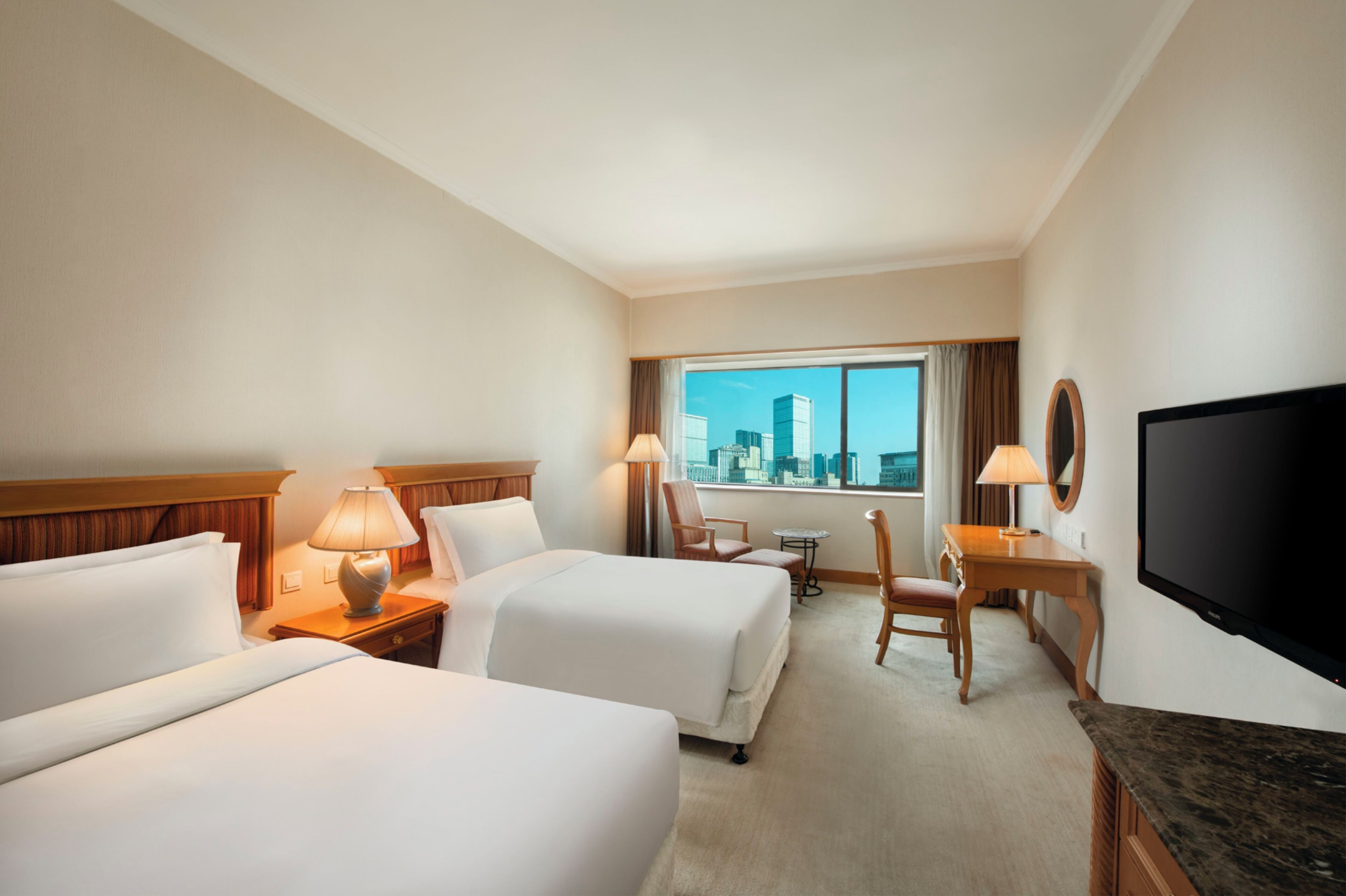 A hotel room with two beds, a desk, a chair, a TV, and a window with a city view.