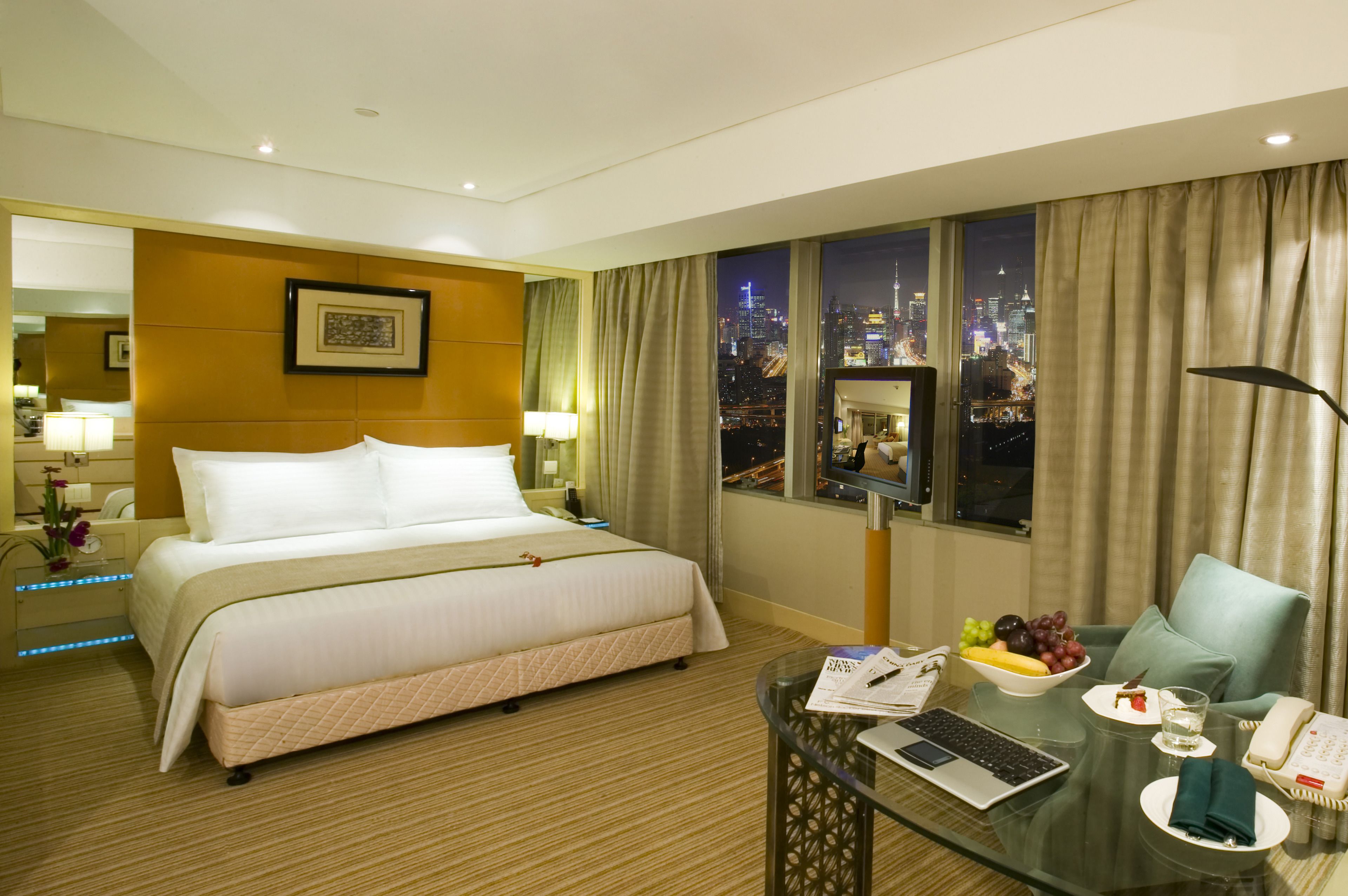 Habitación de hotel con una cama grande, un escritorio con una computadora portátil, un frutero y vistas a un paisaje urbano nocturno.