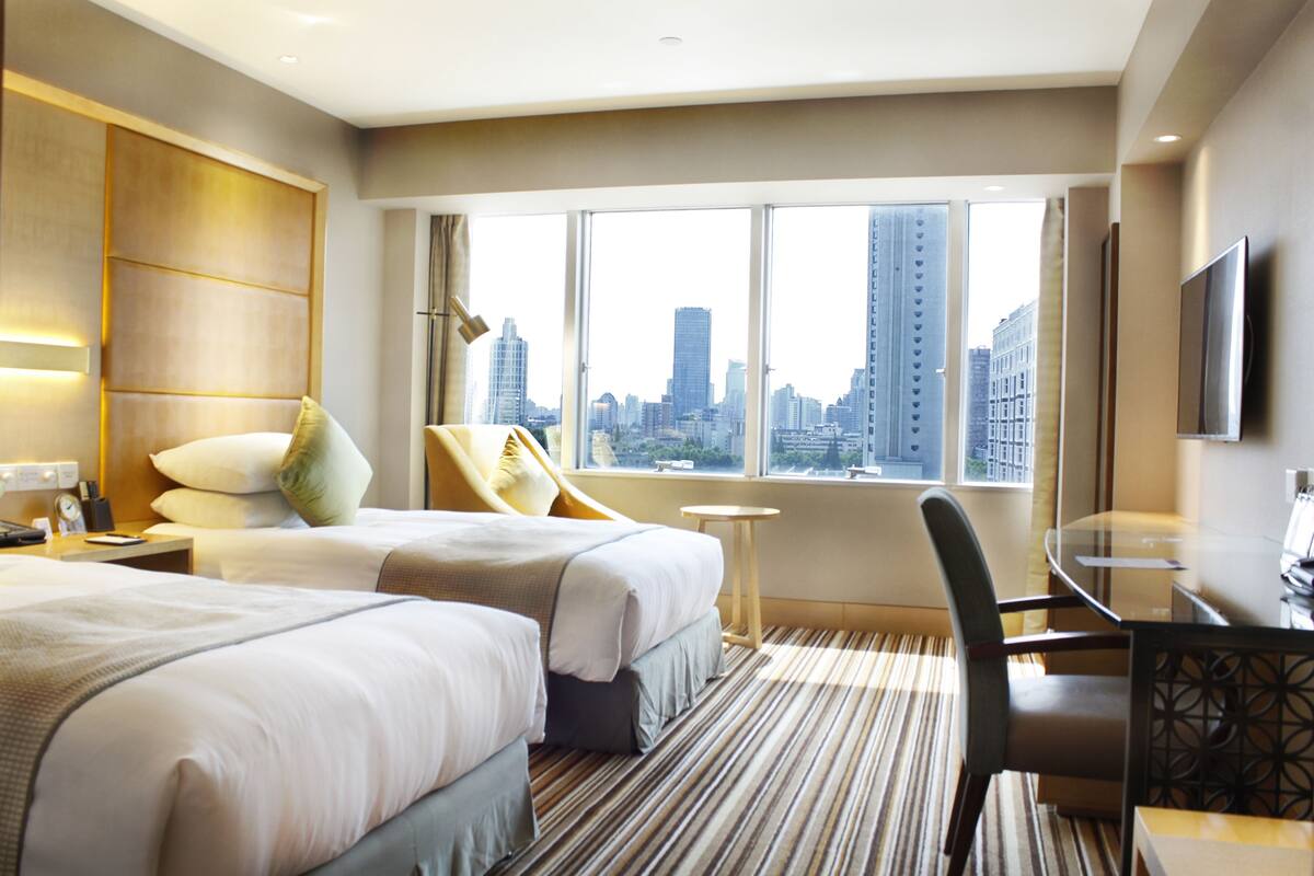 A hotel room with two beds, a desk, a chair, and a large window offering a city view.