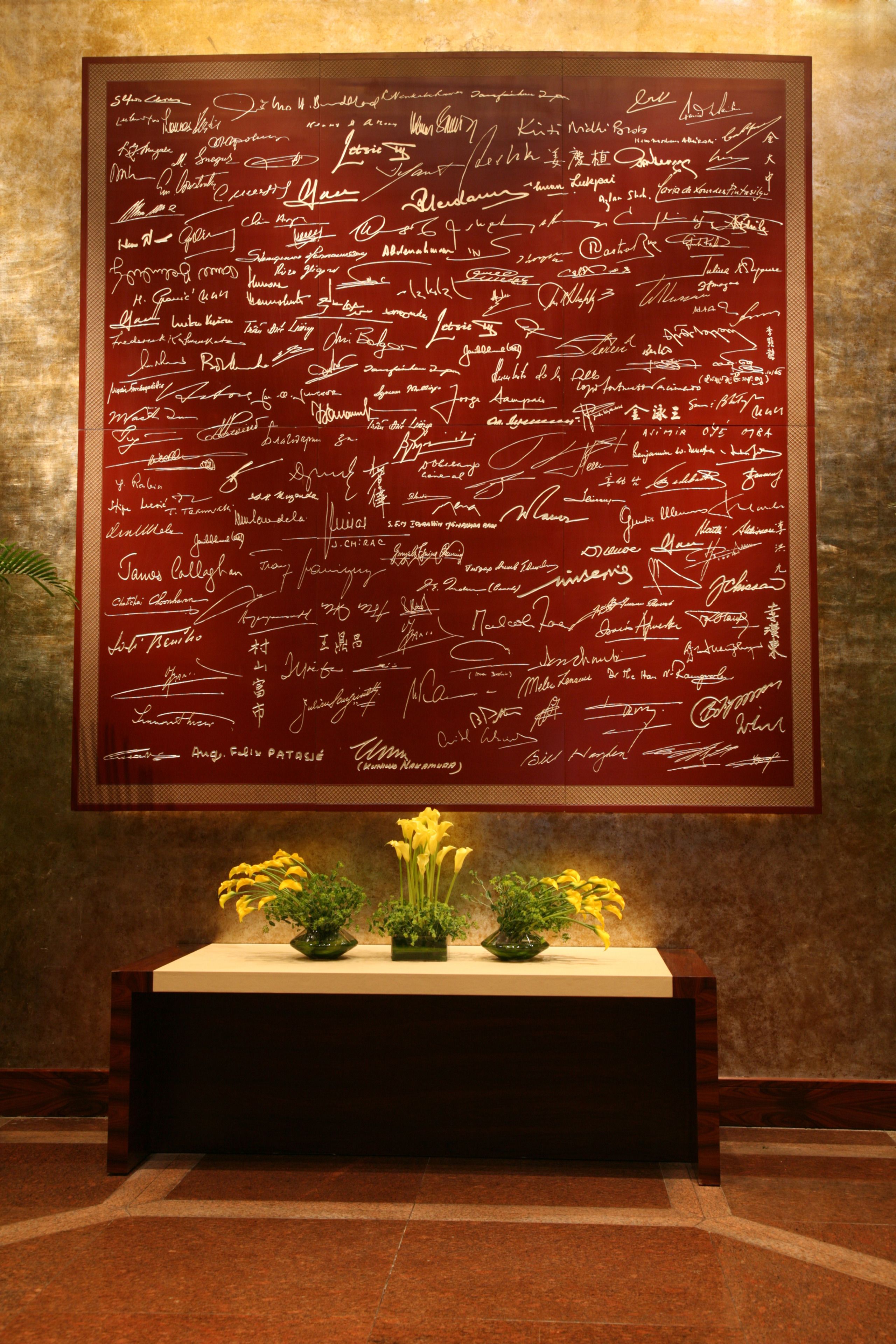 A large red wall with numerous signatures. Below the wall, there is a wooden bench with a vase of yellow flowers.