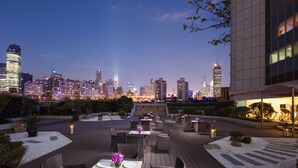 Un área de comedor al aire libre con mesas y sillas, rodeada de guijarros blancos y plantas, con vistas a un horizonte urbano al atardecer.