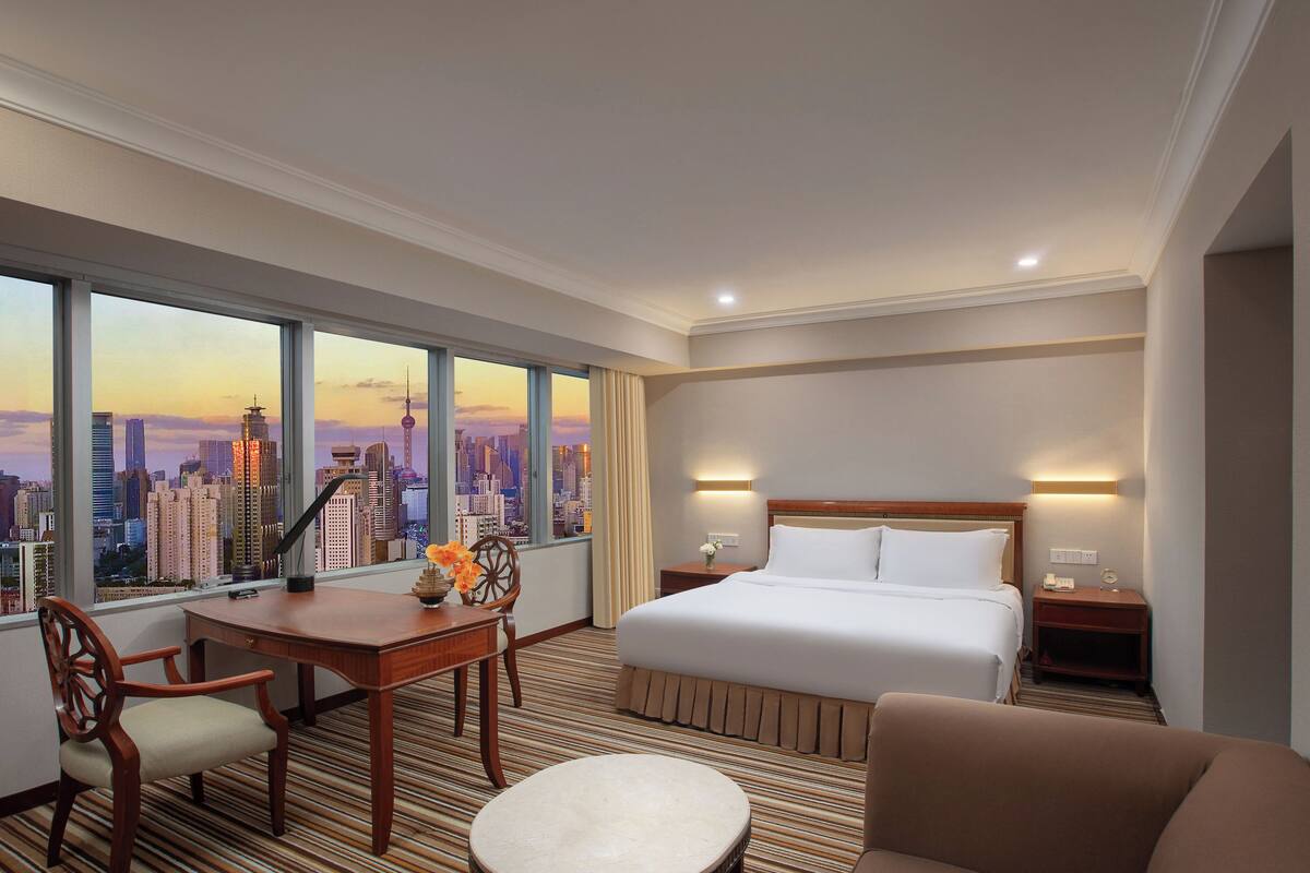 A hotel room with a large window offering a city view, a bed with white linens, a desk with a chair, and a small table with a vase of flowers.