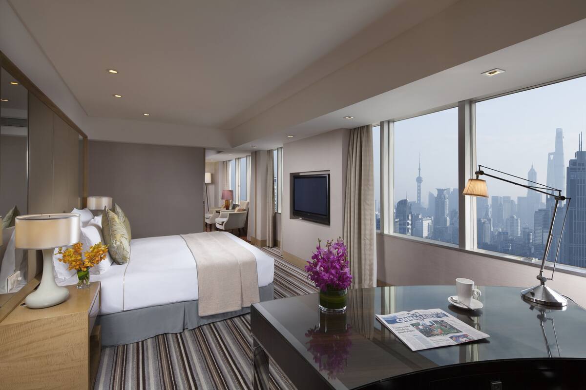 A modern hotel room with a large bed, a desk with a lamp, a vase with flowers, and a view of the city skyline.
