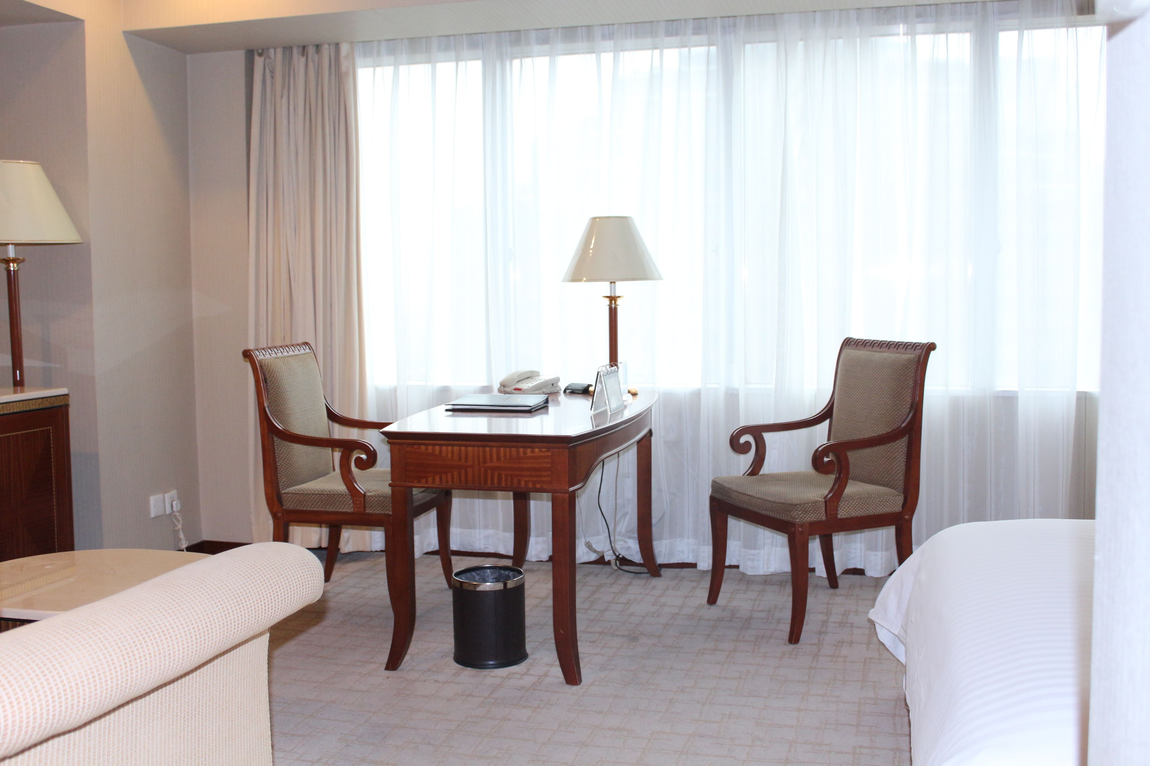 Una habitación de hotel con dos sillas de madera, una mesita con una lámpara, un teléfono y un cubo de basura.