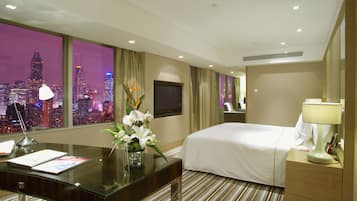A hotel room with a large window offering a city view, a bed, a desk with a lamp, and a mirror.