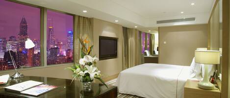 A hotel room with a large window offering a city view, a bed, a desk with a lamp, and a mirror.
