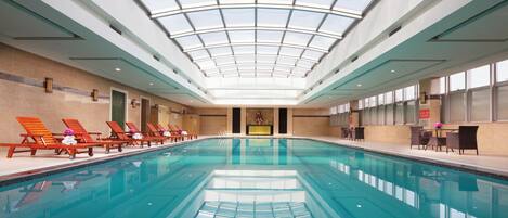 An indoor swimming pool with a glass ceiling, lounge chairs, and a reception area.