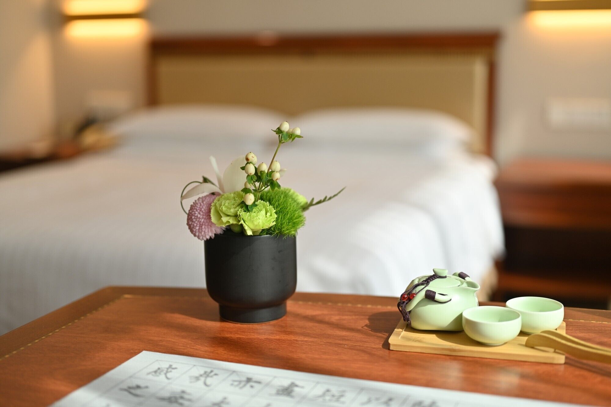 Une chambre d’hôtel bien rangée, avec un lit, une table de chevet agrémentée d’un arrangement floral, une théière et deux tasses.