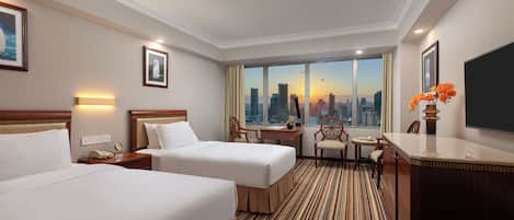 A hotel room with two beds, a desk, a chair, a TV, and a large window with a city view.