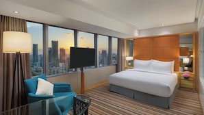 Habitación de hotel con una cama grande, un televisor, un área de descanso con una silla, una mesita de noche con lámpara y vistas a la ciudad a través de amplias ventanas.