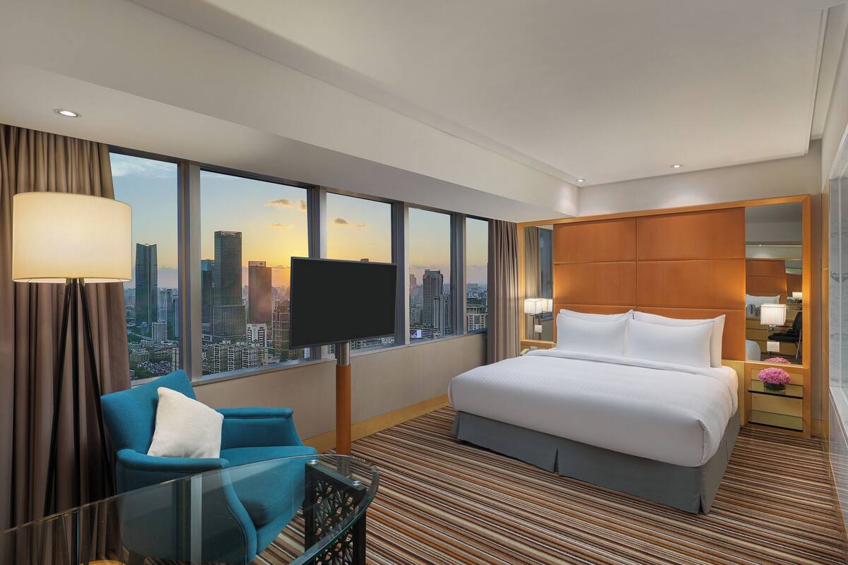 A hotel room with a large bed, a TV, a seating area with a chair, a nightstand with a lamp, and a city view through large windows.