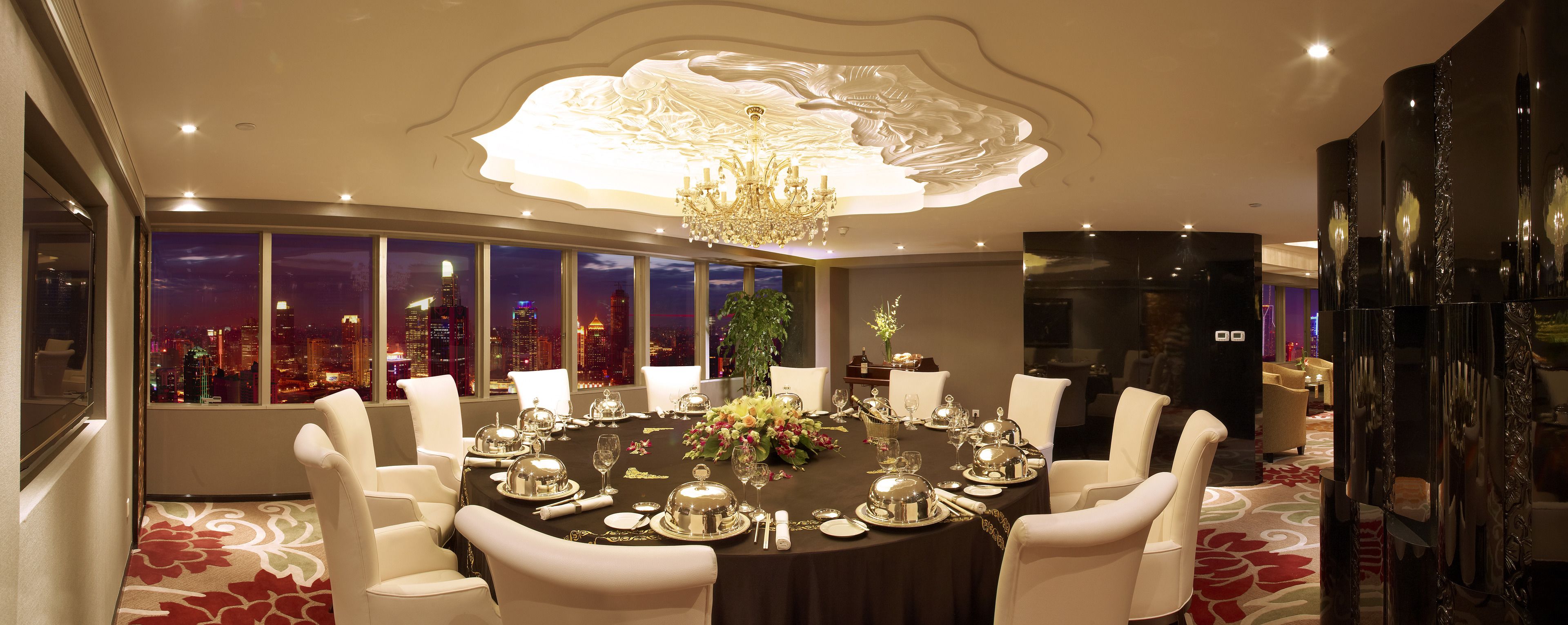 A luxurious dining room with a round table set for six, surrounded by white chairs. A large chandelier hangs from the ceiling, and the room features a large window with a cityscape view.