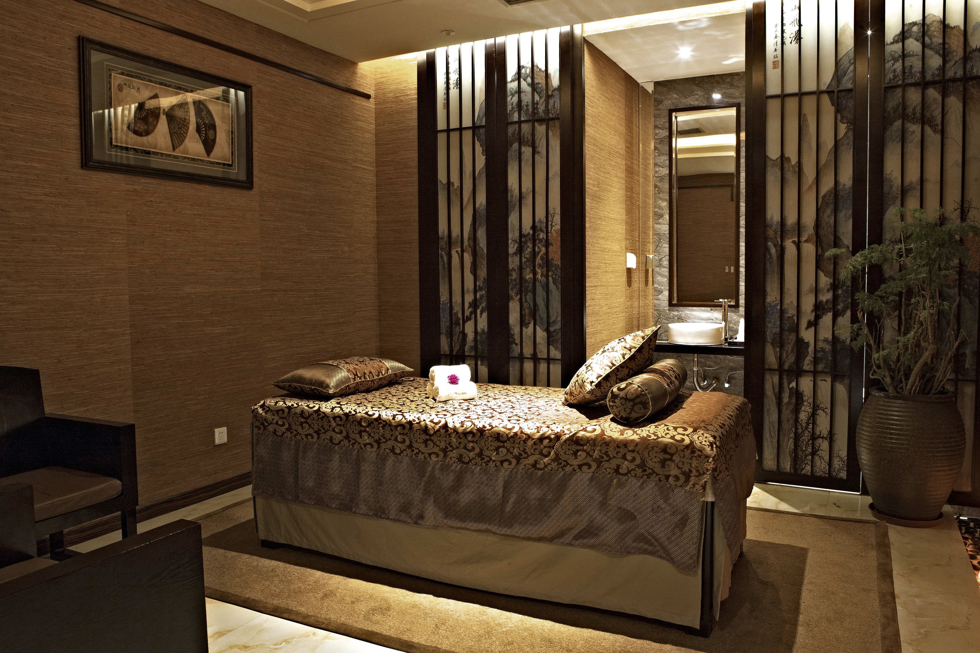 A bedroom with a bed, a chair, a large vase, and a painting on the wall.