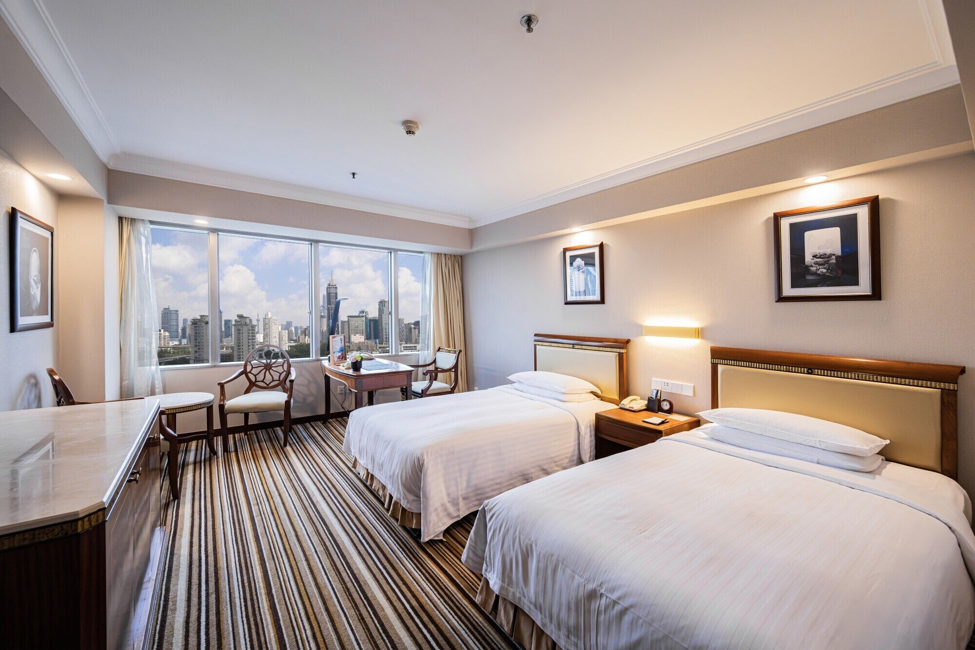 A hotel room with two beds, a desk, a chair, and a view of the city skyline.