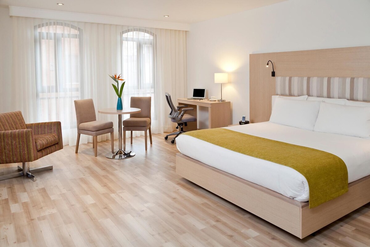 Une chambre d’hôtel avec un grand lit, un bureau avec un ordinateur, deux chaises et une petite table sur laquelle se trouve un vase.