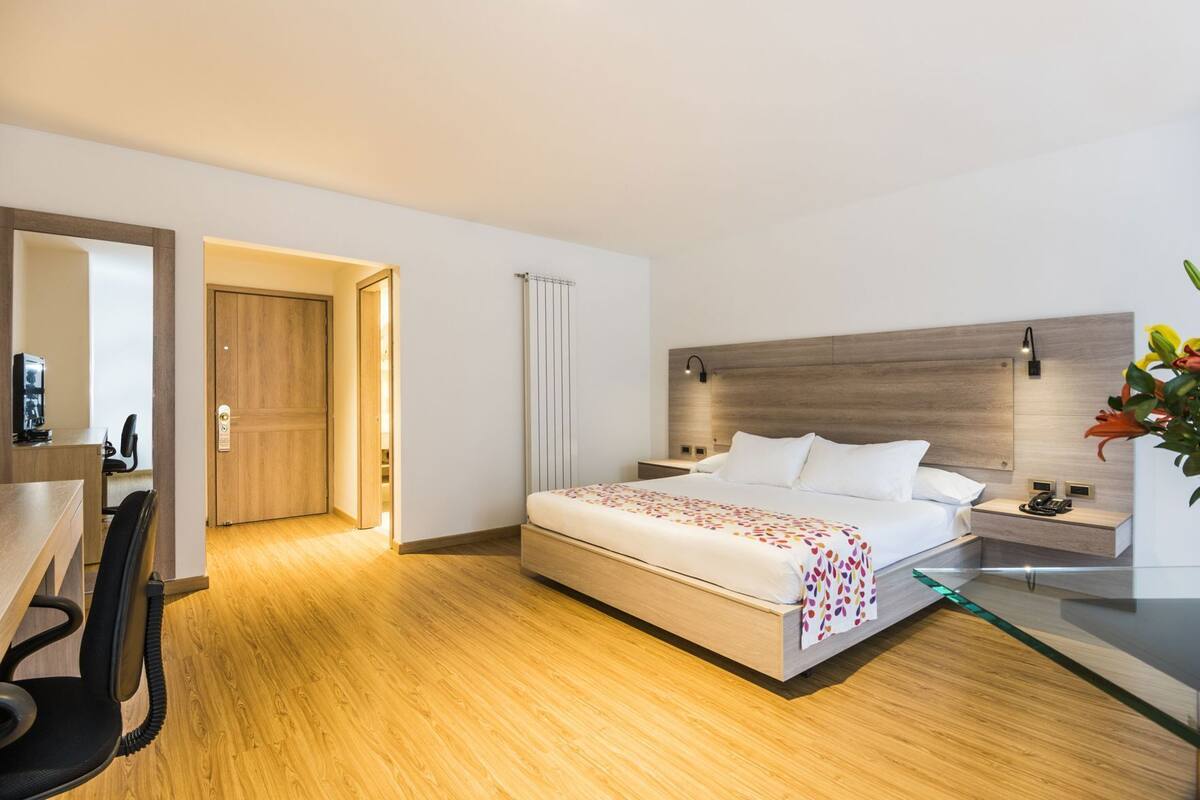 Une chambre d’hôtel avec un plancher en bois, un lit recouvert d’une courtepointe blanche et colorée, un bureau avec une chaise, une table de chevet et une lampe fixée au mur.