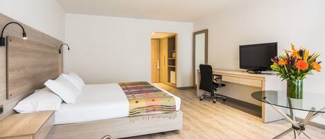 A hotel room with a bed, bedside table, desk, chair, TV, and a glass table with a vase of flowers.