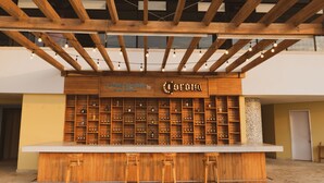 A bar with a wooden counter and stools, shelves with bottles, and a sign reading "Sandbar by Corona". - Las Americas Casa de Playa (Cartagena)