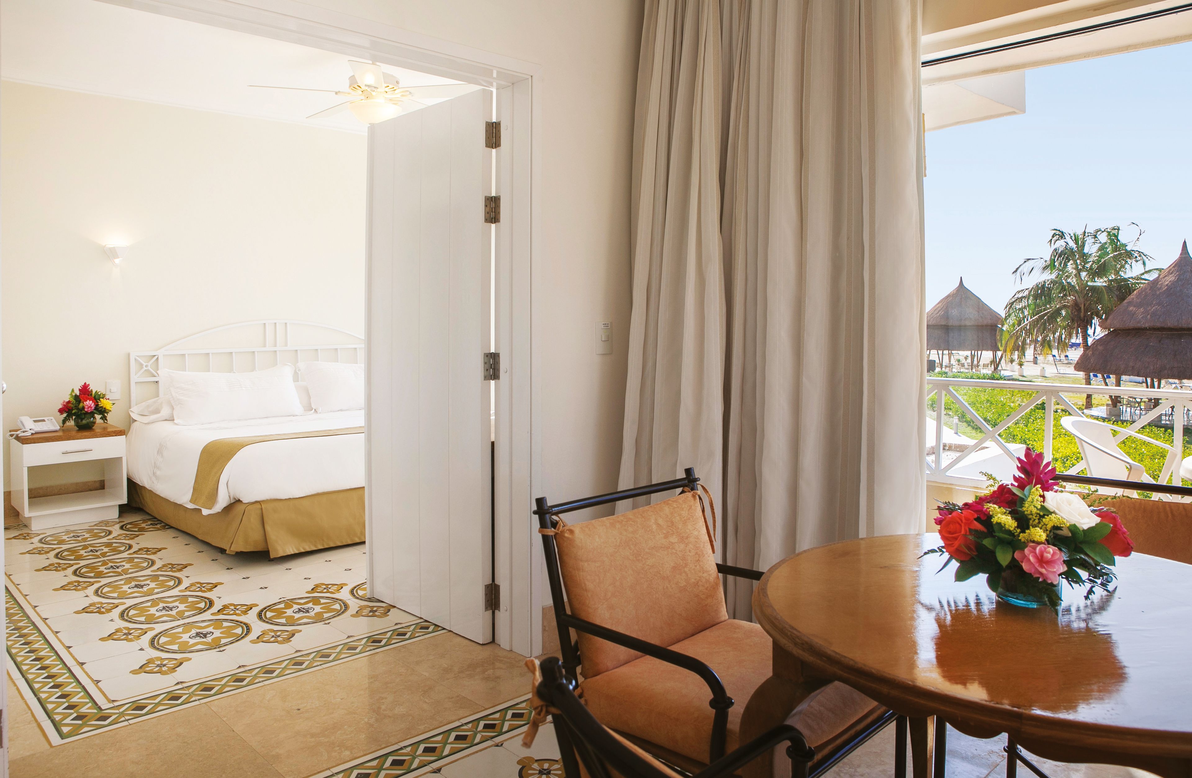 A hotel room with a bed, a chair, a table with a vase of flowers, and a view of a resort with palm trees and thatched-roof huts.
